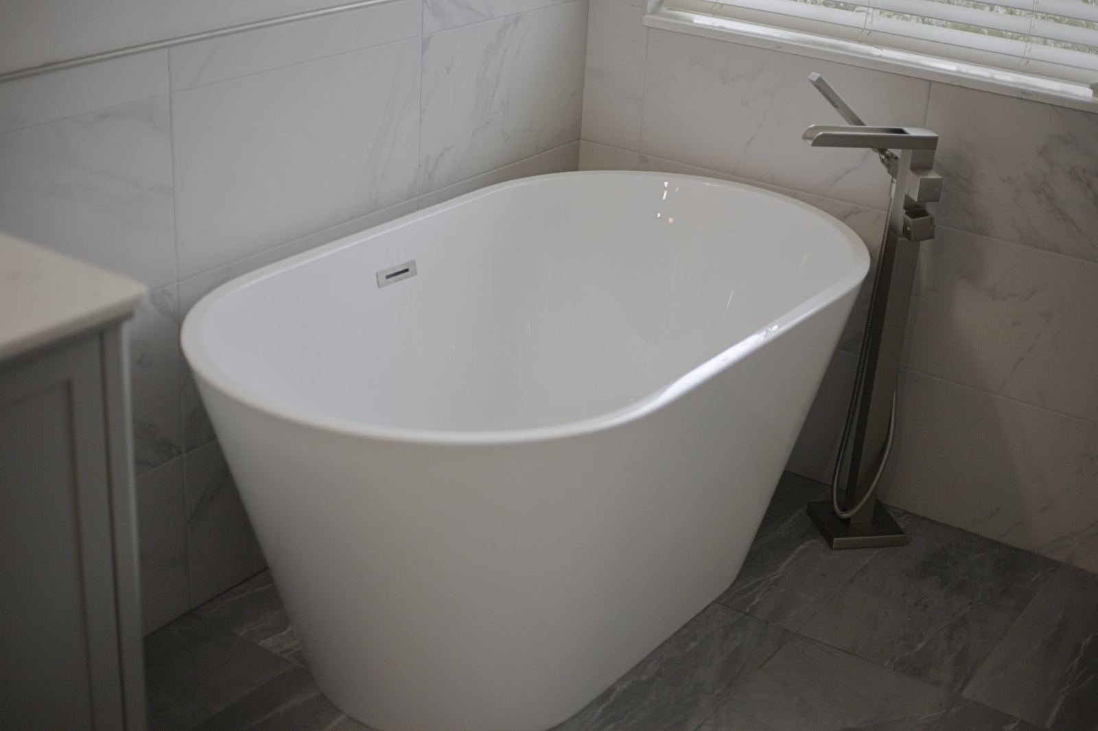 White freestanding tub with a brushed-nickel floor-mounted filler, set against marble-look wall tile on a gray stone-look floor