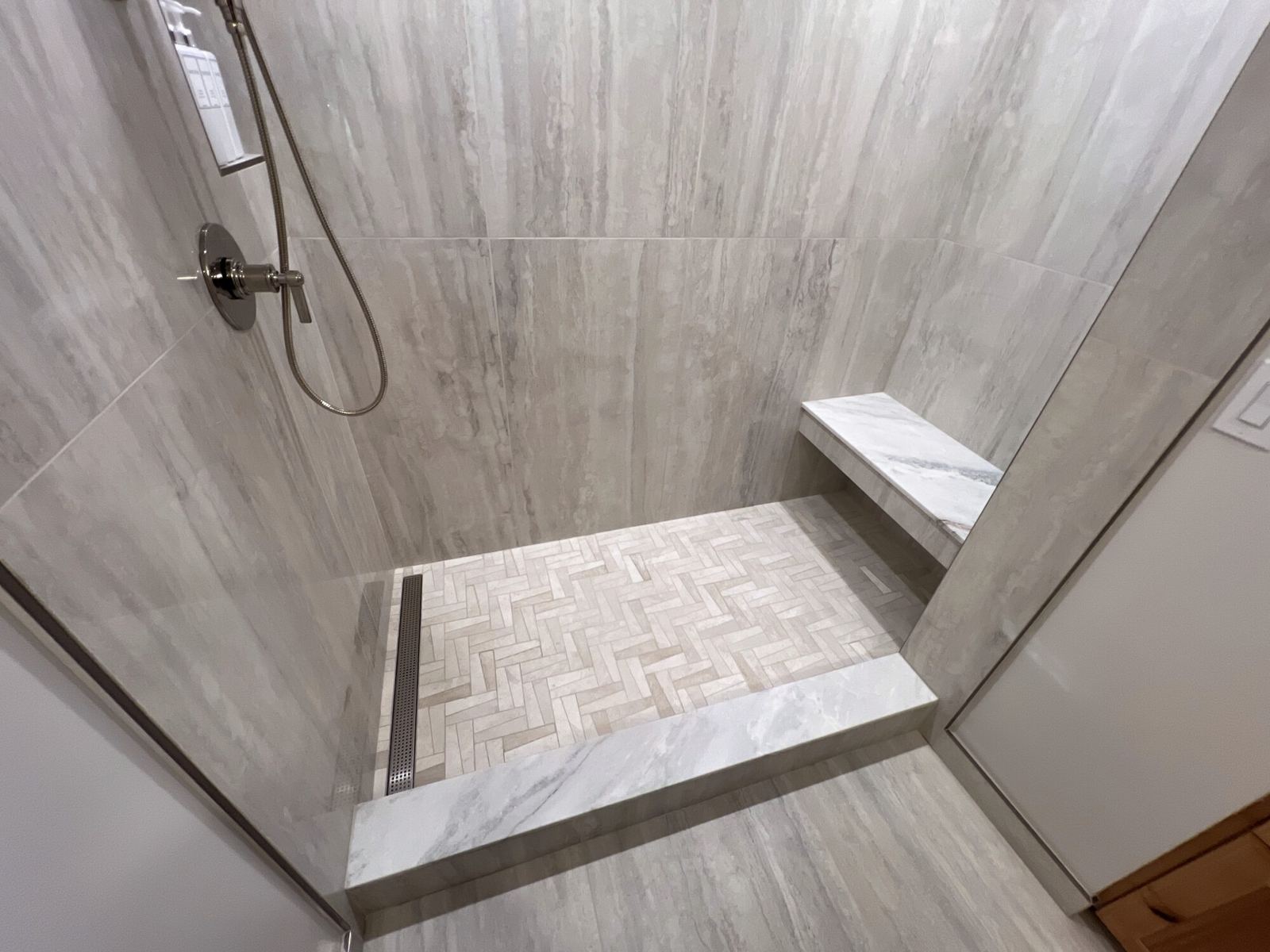 Shower floor in a herringbone pattern with a linear drain, a corner bench and a stone-capped curb