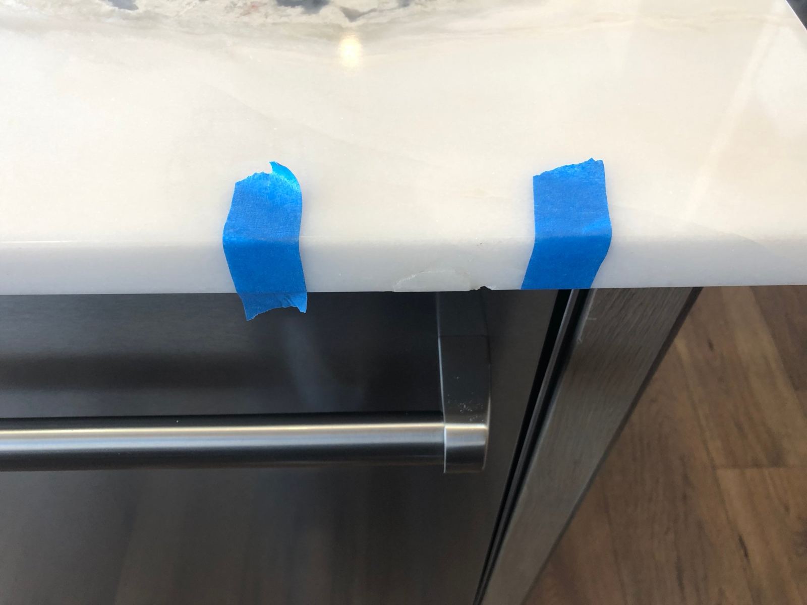 A chip in the edge of a white stone countertop above a dishwasher, marked on each side with blue painter's tape